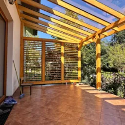 Zadaszenie tarasu z drewna i szkła, widoczne drewniane żaluzje i płytki podłogowe. Słoneczny dzień, widok na ogród.