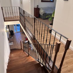Ontera Design & Build - Widok z góry na brązowe schody z czarną metalową balustradą i drewnianymi elementami. W tle widoczny salon z sofą i kominkiem.