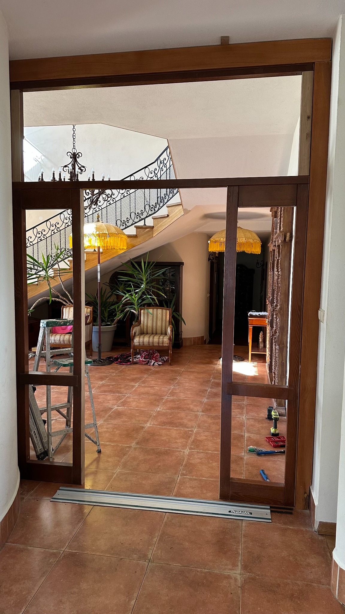 Montaż drewnianych drzwi przesuwnych w domu z terakotową podłogą. Widoczne narzędzia i schody z kutą balustradą. Praca w toku.