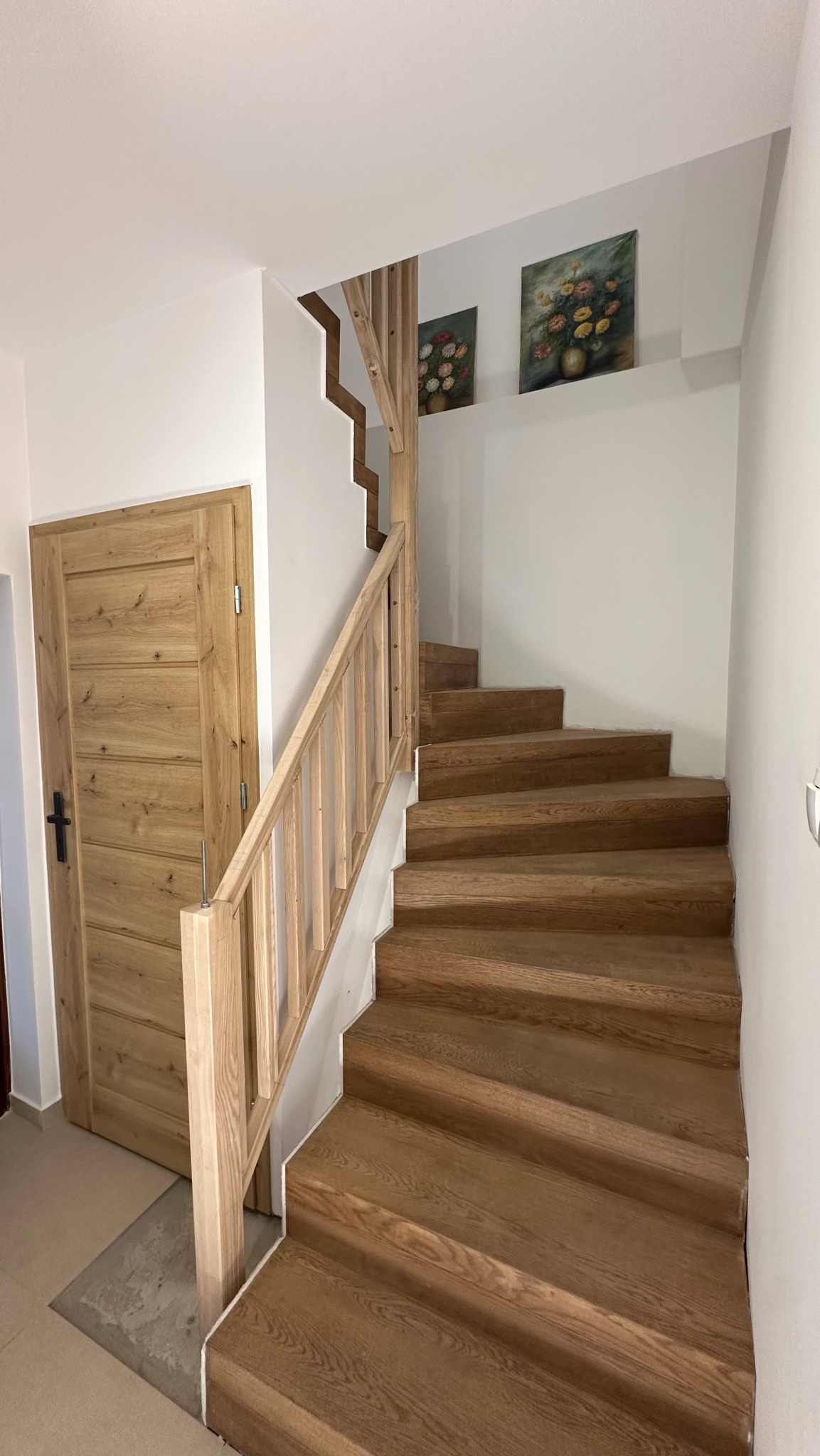 Jasne schody drewniane z balustradą i dopasowane drzwi w minimalistycznym wnętrzu. Uwagę przykuwają obrazy z motywem kwiatowym nad schodami.