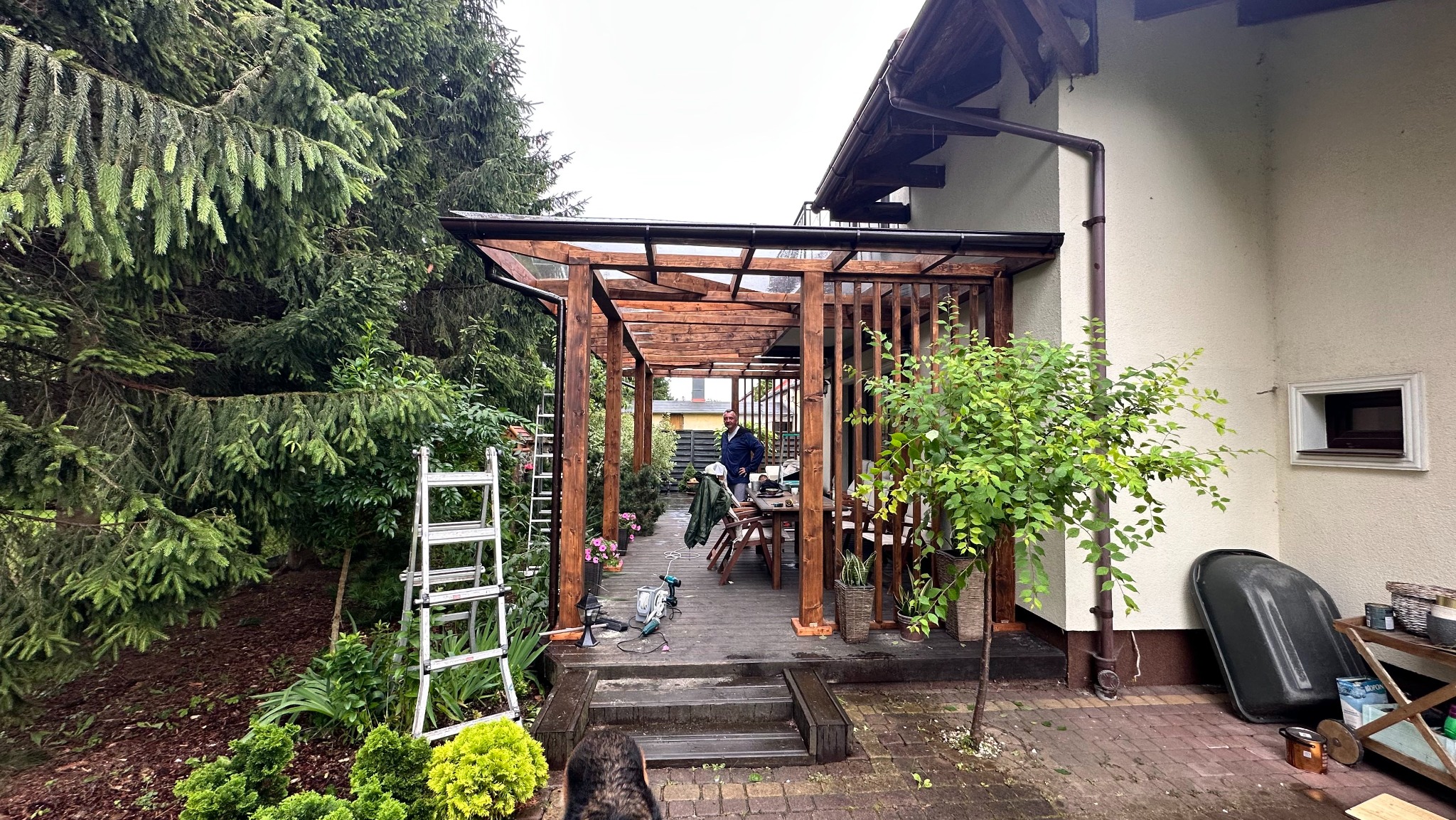 Drewniana pergola z zadaszeniem z poliwęglanu, przylegająca do domu, otoczona zielenią. Widoczne narzędzia i częściowo sylwetka osoby w tle, sugerujące etap budowy.