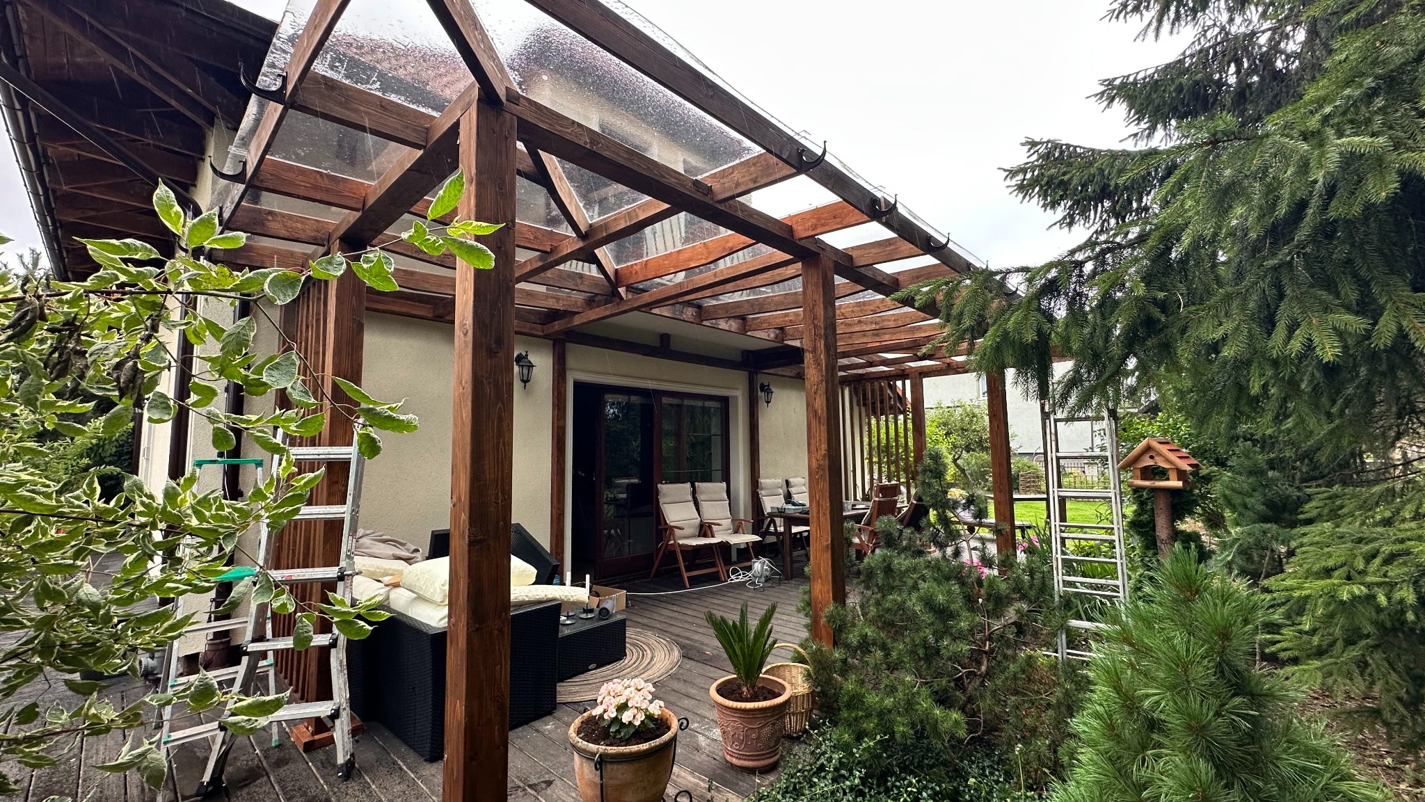 Drewniana pergola z przezroczystym zadaszeniem, meble ogrodowe na tarasie, drabina i roślinność. Widok na dom z zewnątrz, relaksacyjna przestrzeń na świeżym powietrzu.