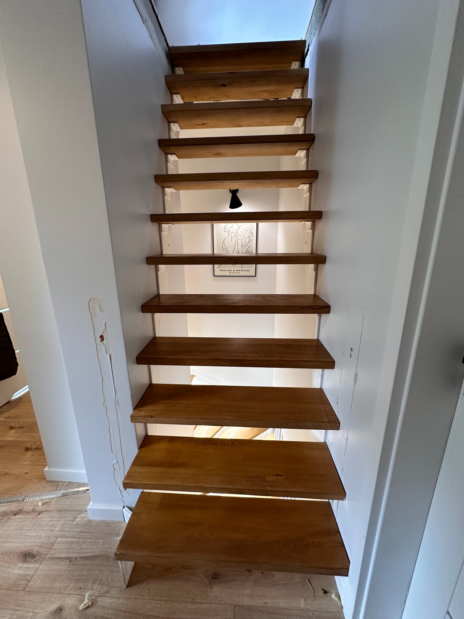 Wąskie schody drewniane w jasnym wnętrzu, widok od dołu. Stopnie oświetlone od spodu, na ścianie grafika w ramce. Nierówności na ścianie po lewej stronie.