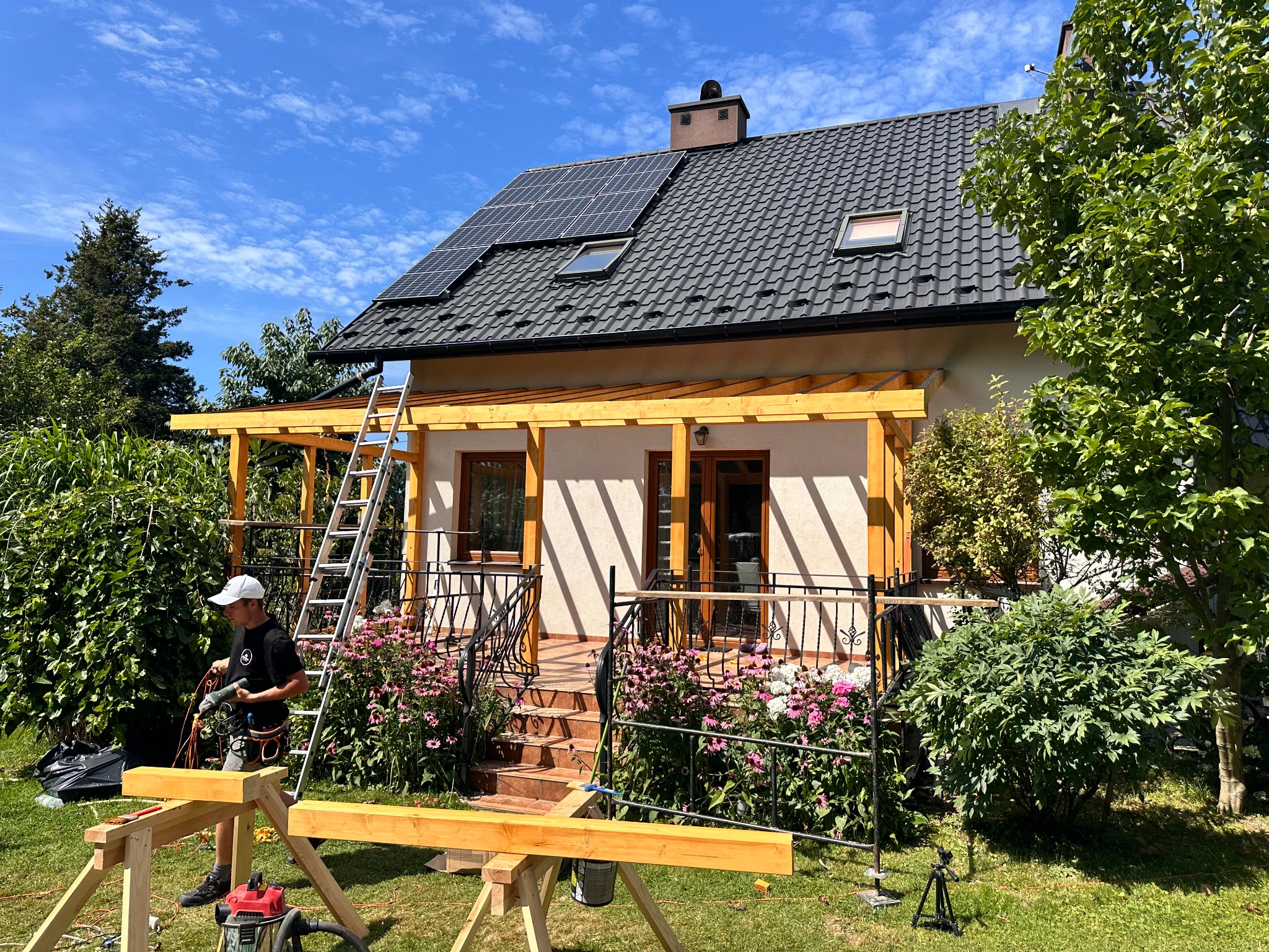 Konstrukcja drewniana werandy przy domu jednorodzinnym w słoneczny dzień. Stolarz przy pracy, widoczne narzędzia i elementy konstrukcyjne.