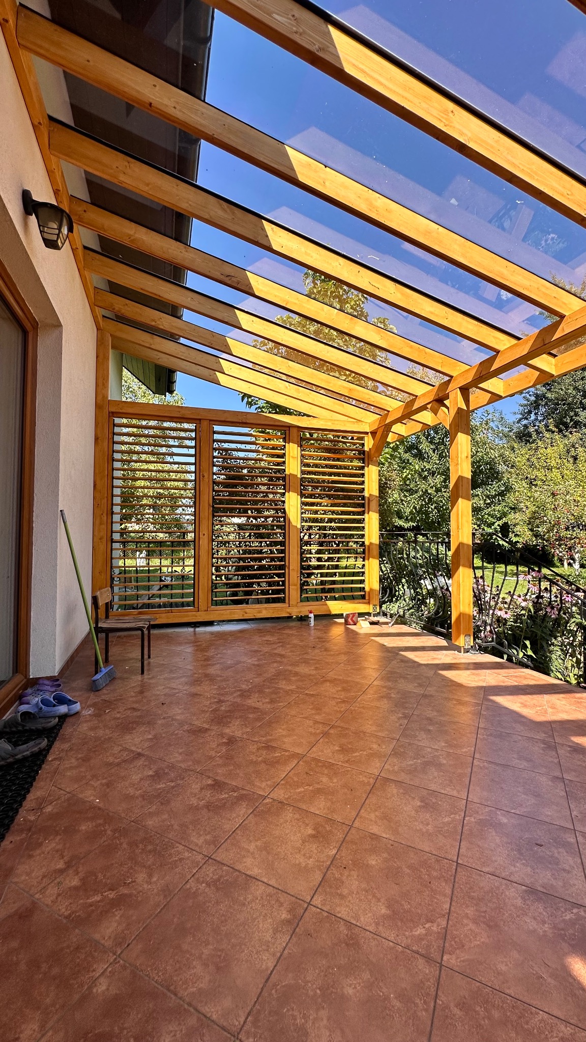 Zadaszenie tarasu z drewna i szkła, widoczne drewniane żaluzje i płytki podłogowe. Słoneczny dzień, widok na ogród.
