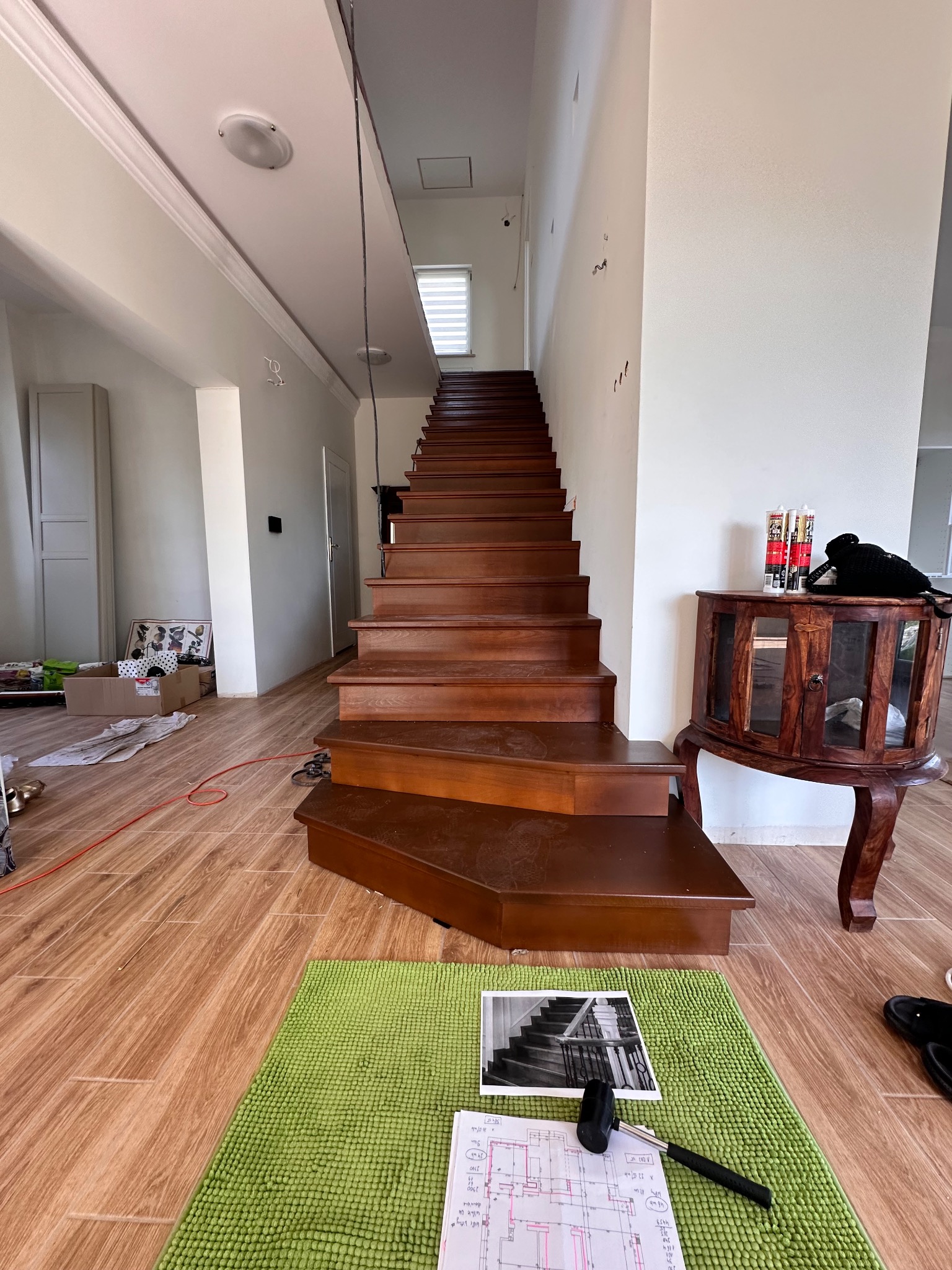 Wnętrze domu z drewnianymi schodami w trakcie renowacji. Na pierwszym planie zielony dywanik z planami i narzędziami. W tle komoda i kartony.