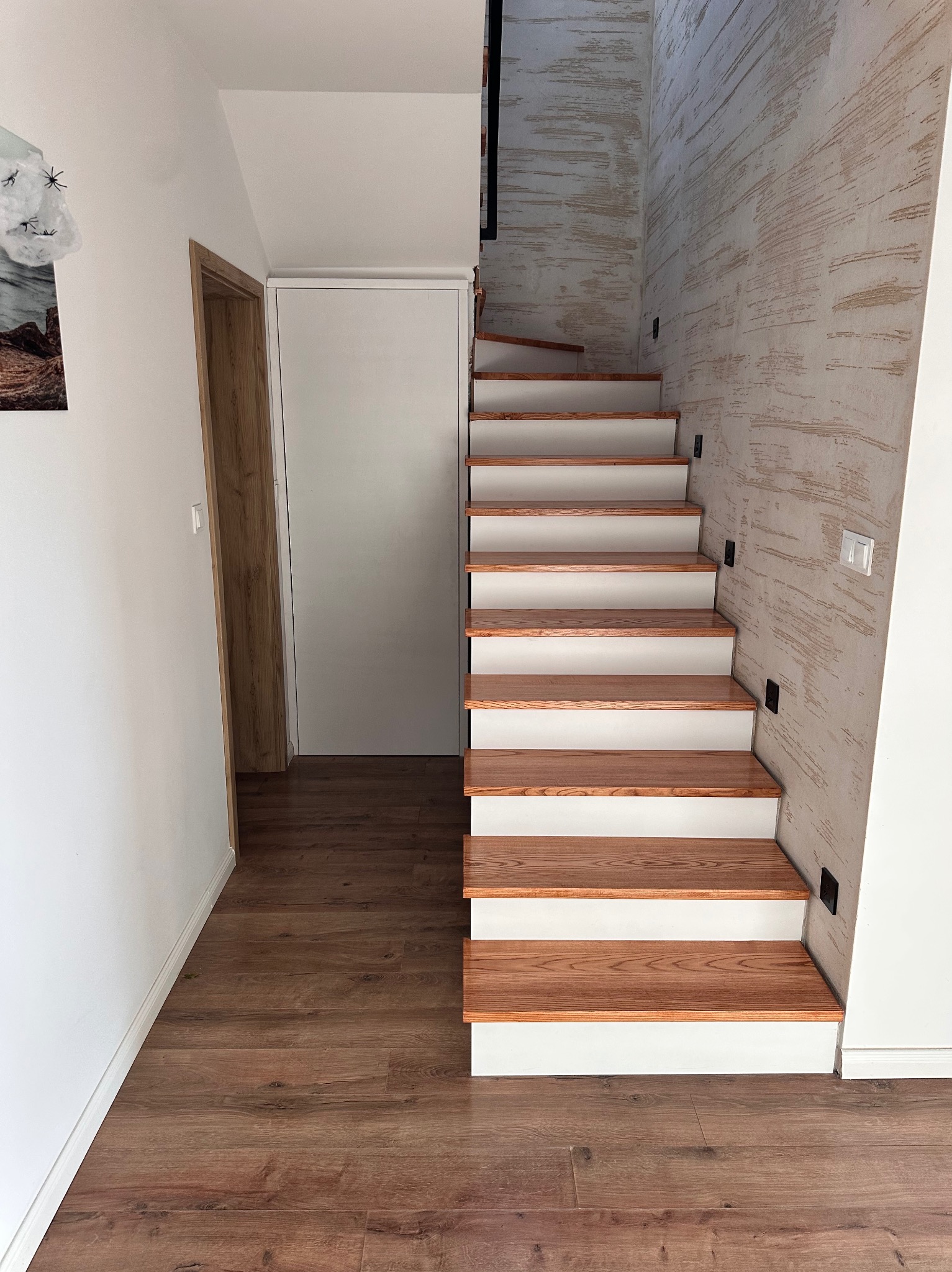 Nowoczesne schody z drewnianymi stopniami i białymi podstopnicami, prowadzące w górę. Ściana z dekoracyjną fakturą. Widoczne drzwi i fragment podłogi z paneli.