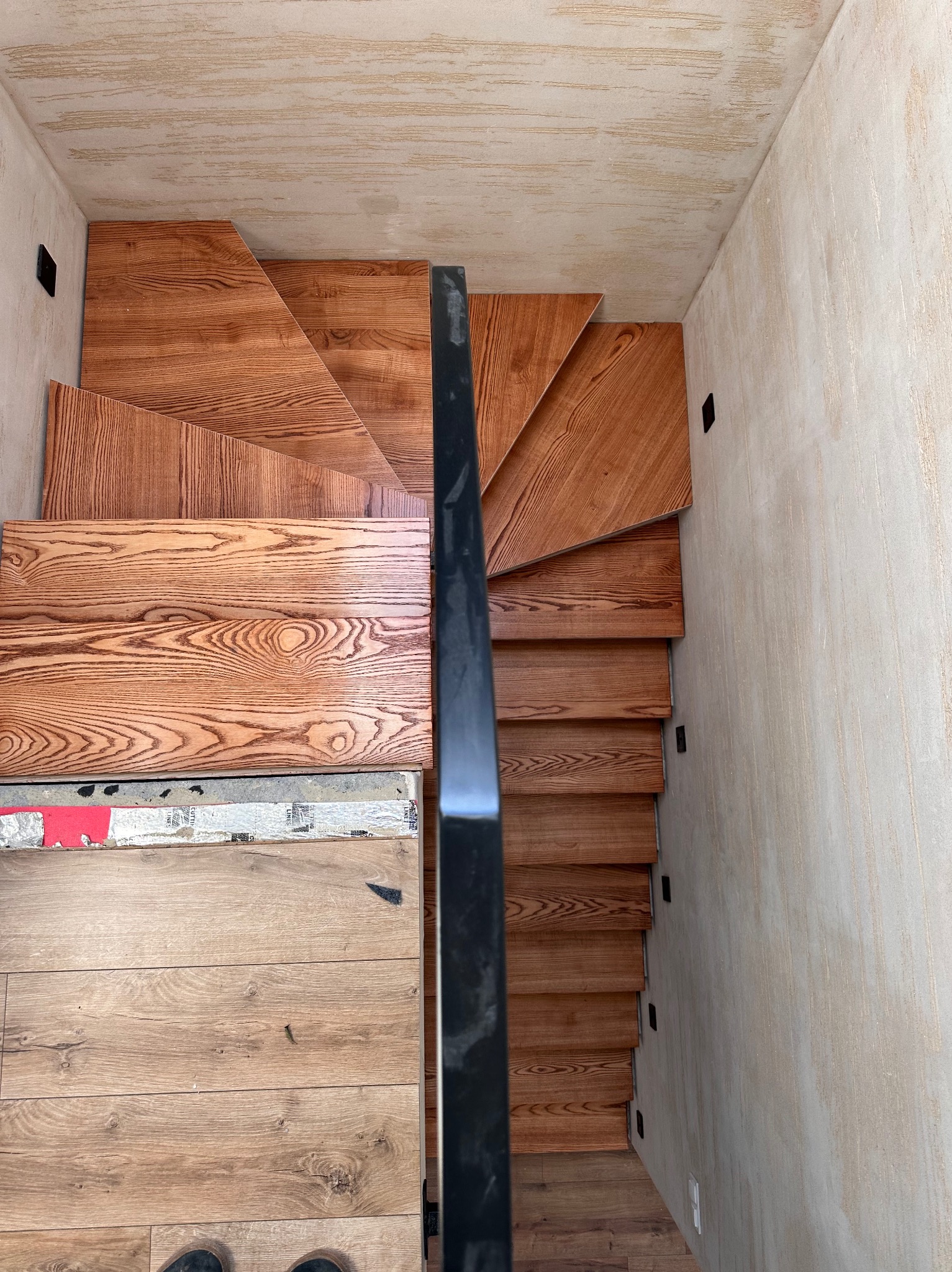 Kątowe ujęcie z góry na drewniane schody z czarną balustradą, widoczne fragmenty podłogi i sufitu. Naturalne oświetlenie podkreśla strukturę drewna.