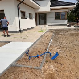 MAKOST BRUKARSTWO - Montaż białej kostki brukowej na posesji domu jednorodzinnego. Widoczne przygotowane podłoże i pracownicy w trakcie pracy. Częściowo ułożona nawierzchnia.