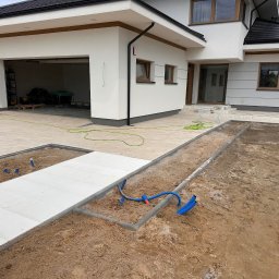 MAKOST BRUKARSTWO - Nowoczesny dom z jasną kostką brukową na podjeździe i elementami instalacji podziemnej w trakcie prac ziemnych. Widoczna ubijarka w tle.