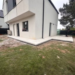 MAKOST BRUKARSTWO - Nowoczesny taras z jasnych płyt przy białym domu. Widoczni dwaj pracownicy podczas czyszczenia powierzchni. Trawa i drzewo w tle.