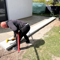 MAKOST BRUKARSTWO - Pracownik z poziomnicą i młotkiem gumowym układa jasnoszare płyty na podwórku, w tle widoczni dwaj mężczyźni i szklarnia.