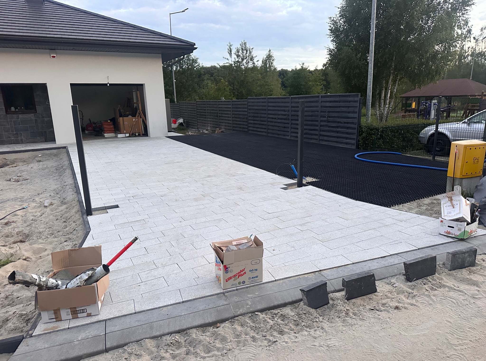 Jasnoszara kostka brukowa ułożona na podjeździe domu jednorodzinnego. Fragment podjazdu wyłożony czarną kratką trawnikową. Widoczne narzędzia i materiały budowlane.