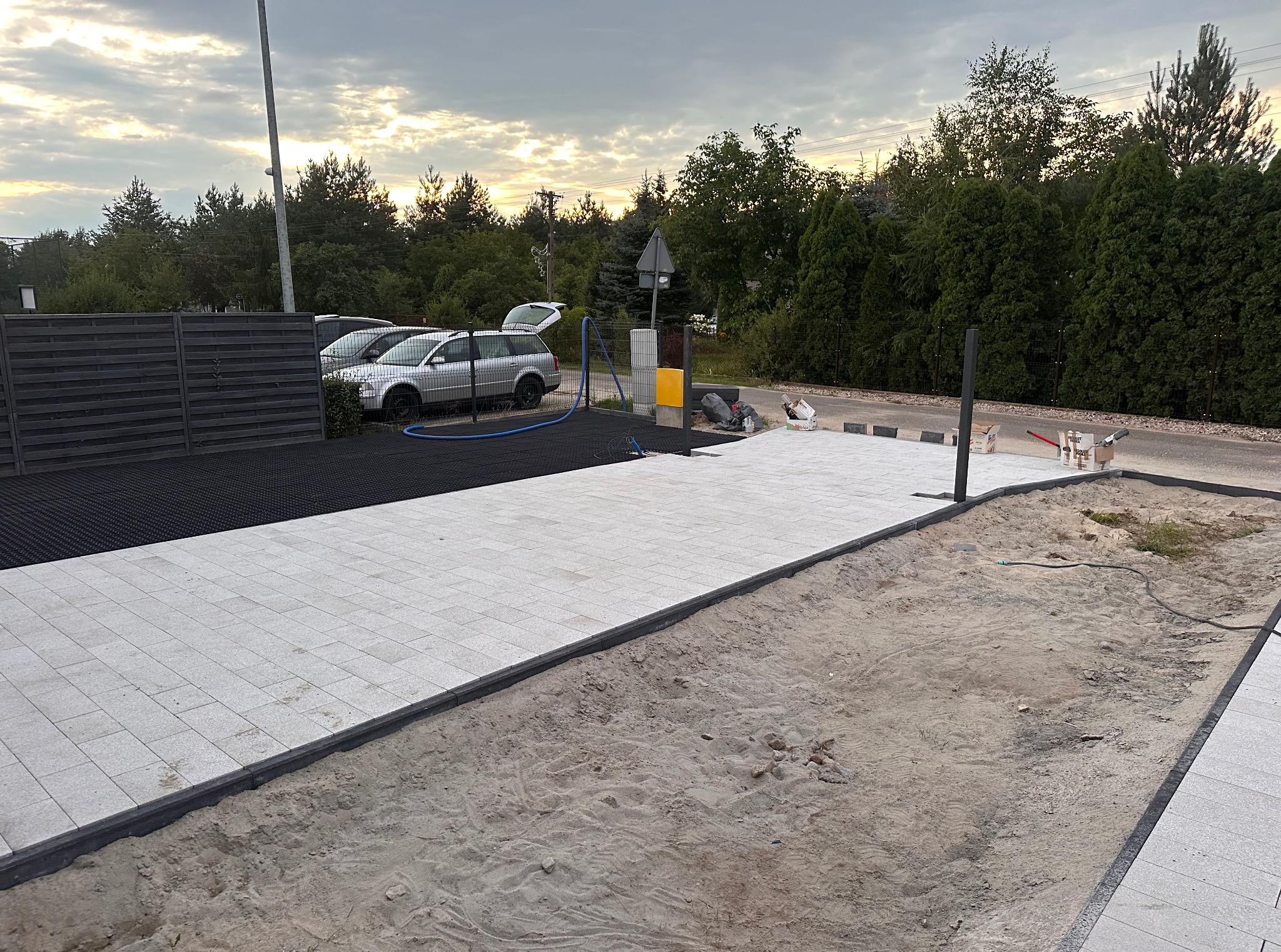 Jasnoszara kostka brukowa z ciemną obwódką, ułożona częściowo na piasku, z widocznym samochodem w tle. Prace brukarskie w toku, widok na posesję z elementami zieleni.