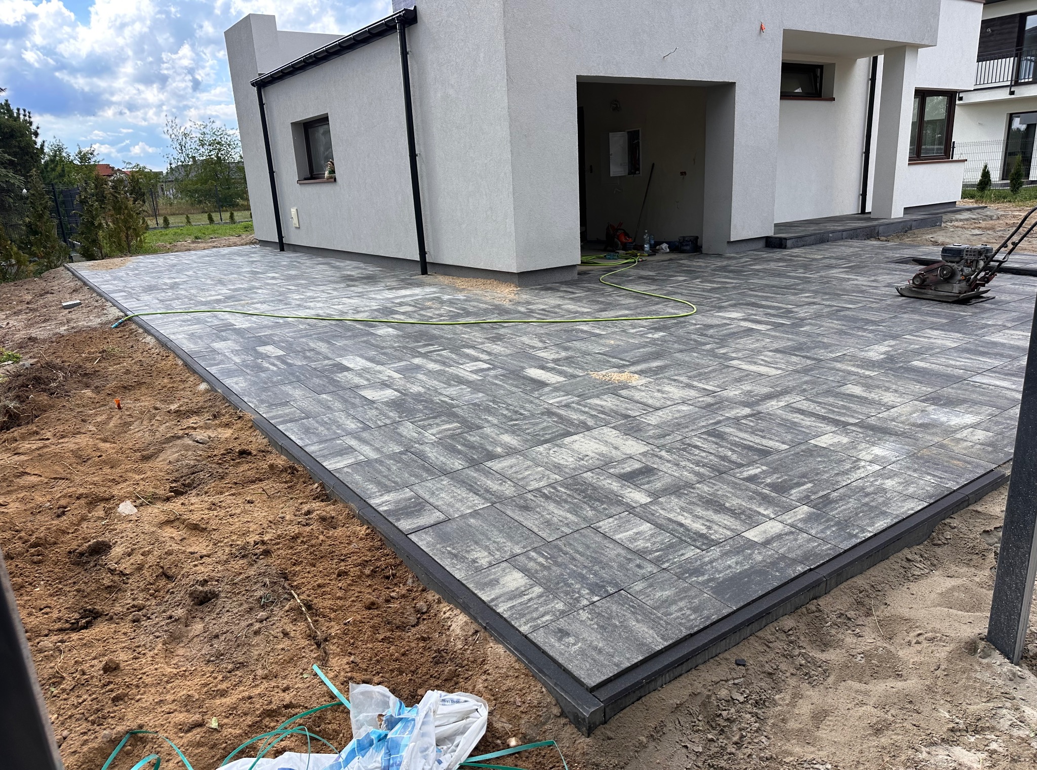 Wykończony taras z szarej kostki brukowej o nieregularnym wzorze, obrzeża w kolorze czarnym. W tle jasny budynek, a na kostce maszyna do ubijania.