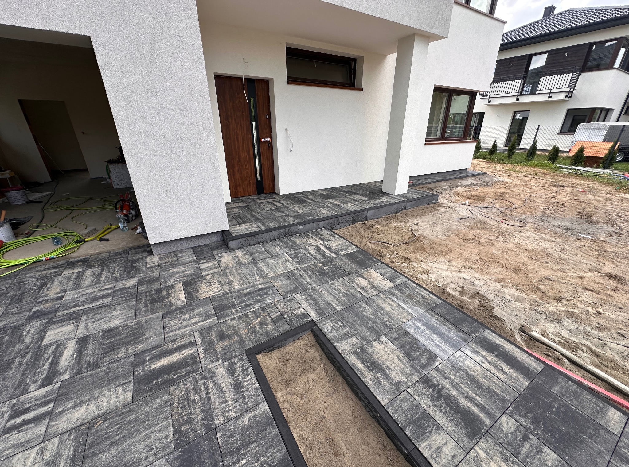 Ułożona, grafitowa kostka brukowa przed nowoczesnym domem. Widoczny fragment wykopu pod dalsze prace. W tle drugi budynek.