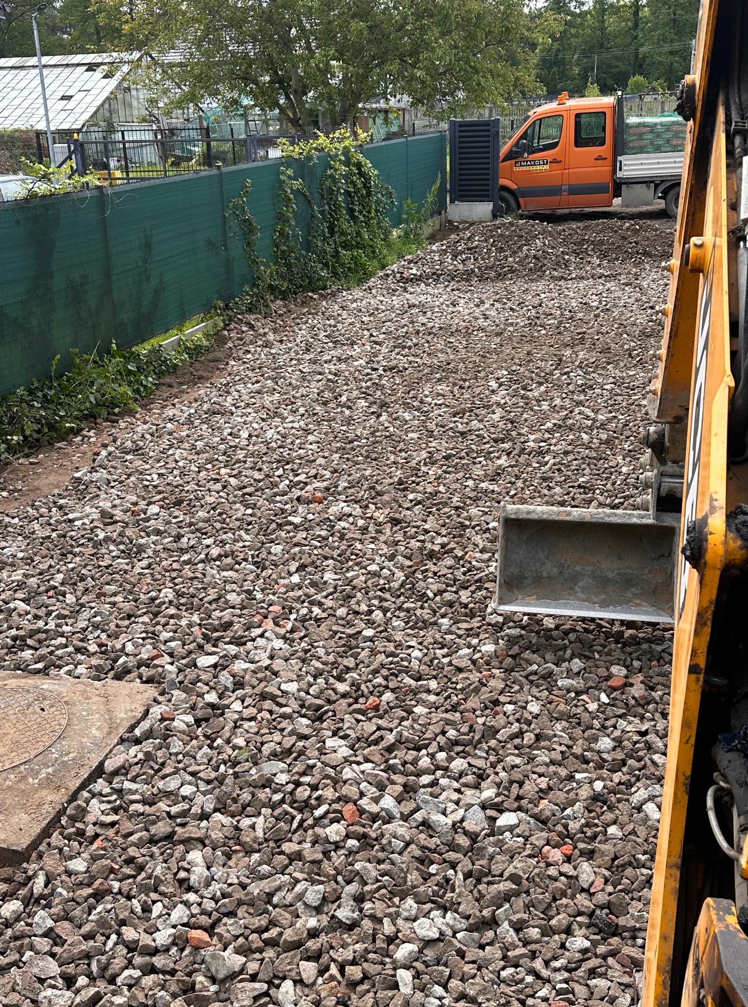 Przygotowany teren pod kostkę brukową z użyciem koparki i dostawczego auta firmy Makost, widok na posesję z zielonym ogrodzeniem.