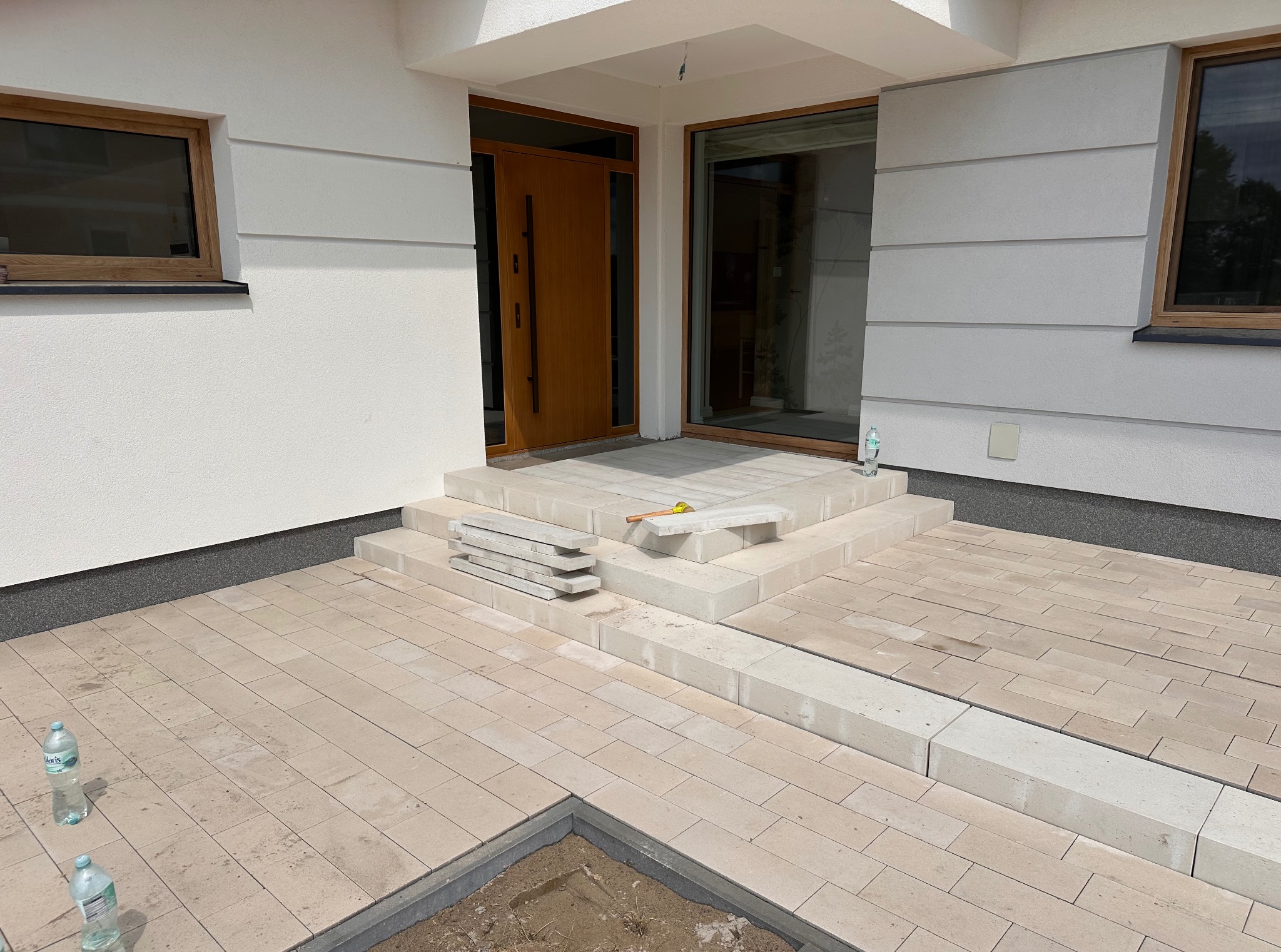 Jasna kostka brukowa ułożona przed nowoczesnym domem. Widoczne schody wejściowe z kostki i fragment niezakończonej nawierzchni. Butelki wody na bruku.