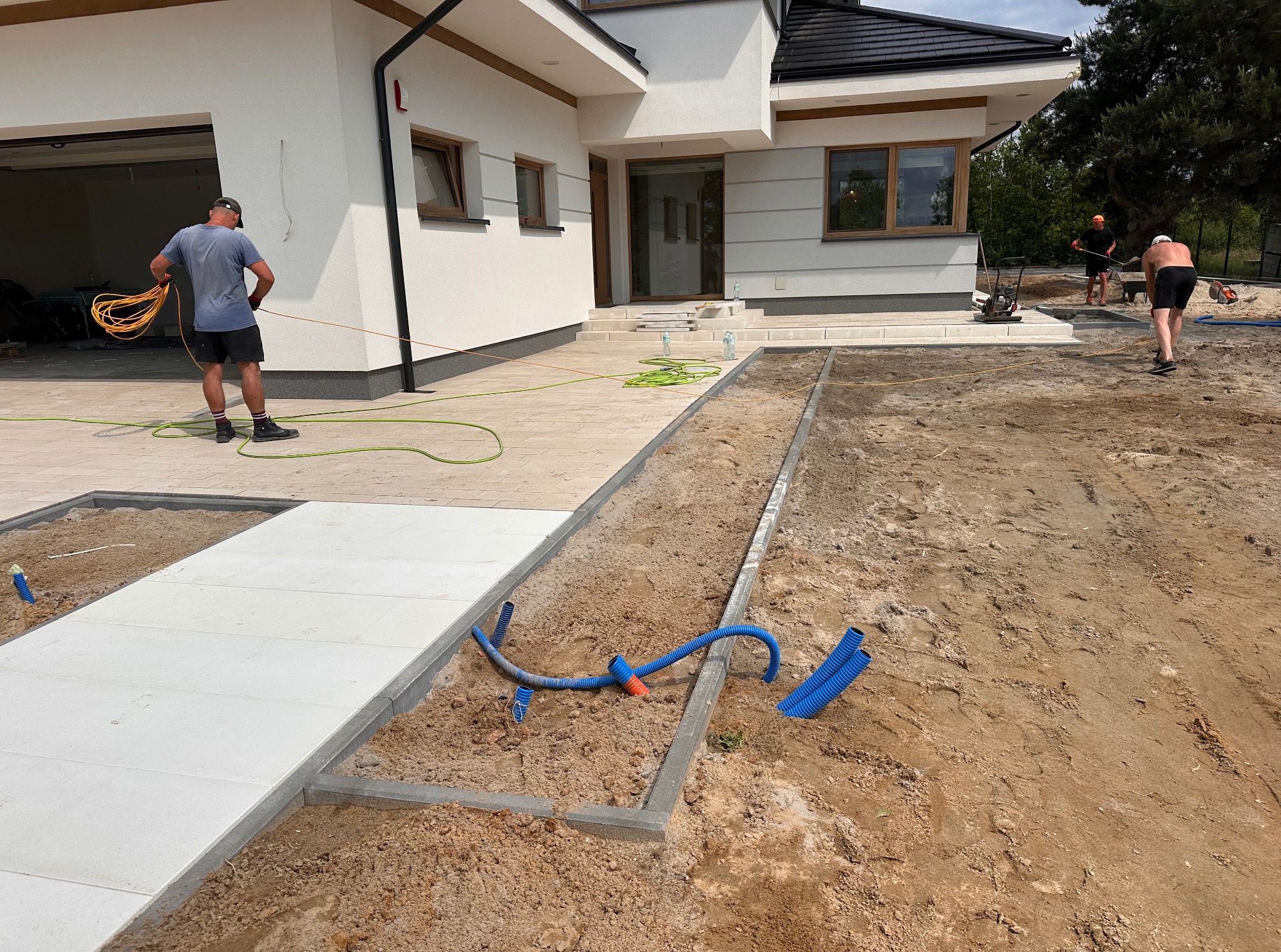 Montaż białej kostki brukowej na posesji domu jednorodzinnego. Widoczne przygotowane podłoże i pracownicy w trakcie pracy. Częściowo ułożona nawierzchnia.