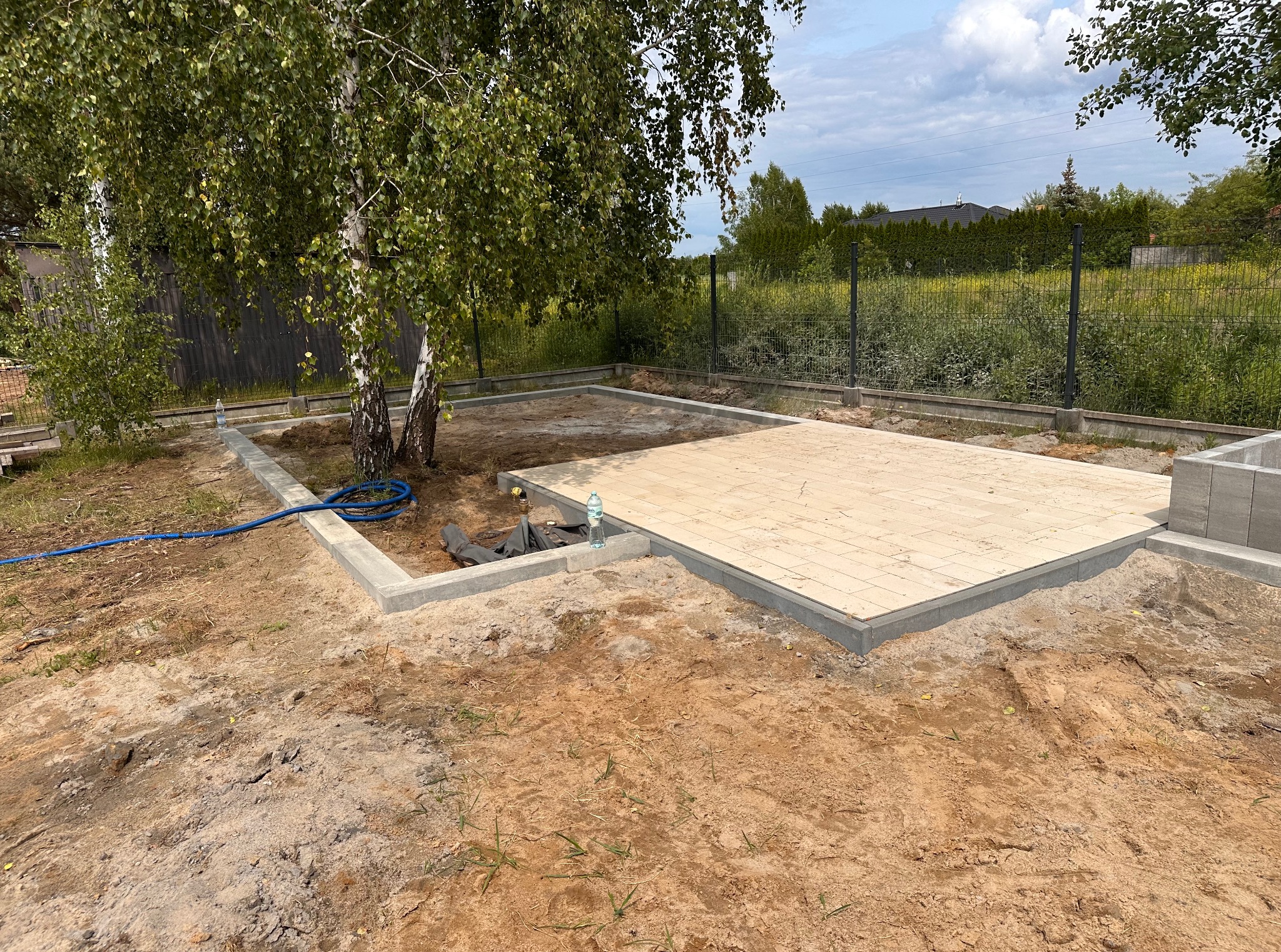 Fragment placu z jasnej kostki, obrzeża z szarego betonu, częściowo otoczony ziemią. W tle drzewa i ogrodzenie z siatki.