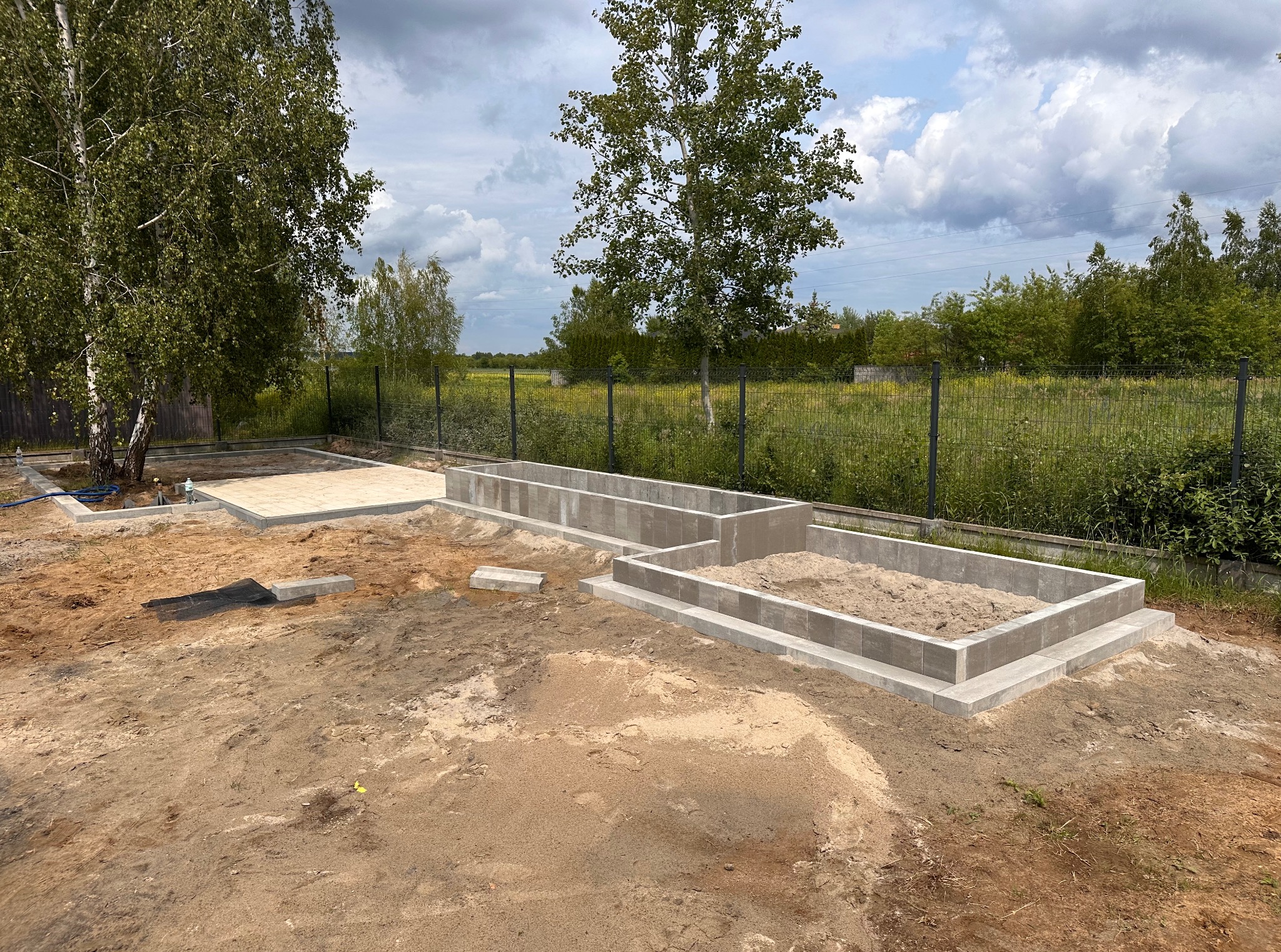 Fundamenty z kostki brukowej w ogrodzie, częściowo wypełnione piaskiem, przygotowane pod budowę, widok z góry na tle zieleni i błękitnego nieba.