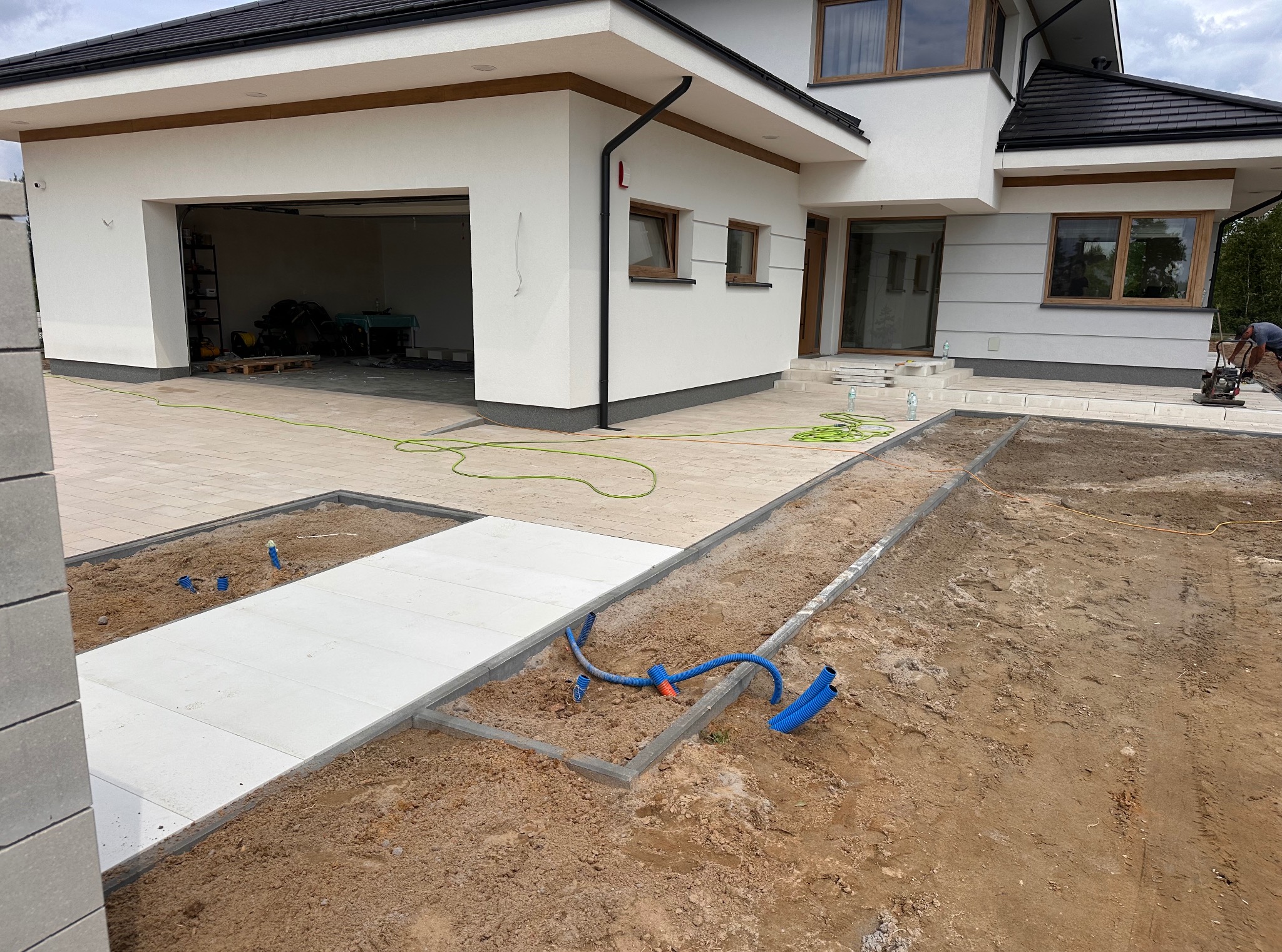 Nowoczesny dom z jasną kostką brukową na podjeździe i elementami instalacji podziemnej w trakcie prac ziemnych. Widoczna ubijarka w tle.