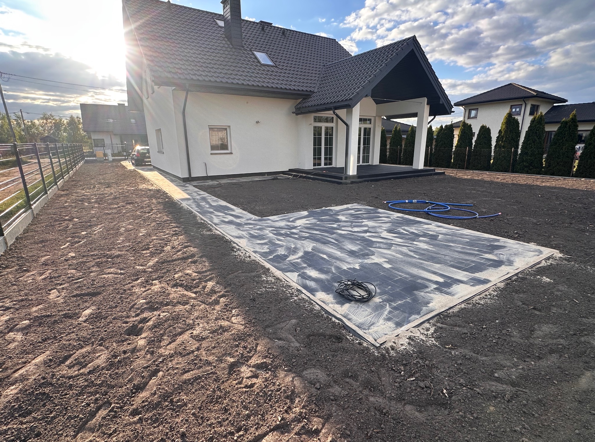 Nowoczesny dom jednorodzinny z częściowo ułożoną kostką brukową na podwórku. Widoczne przygotowane podłoże i materiały budowlane. Słoneczny dzień.