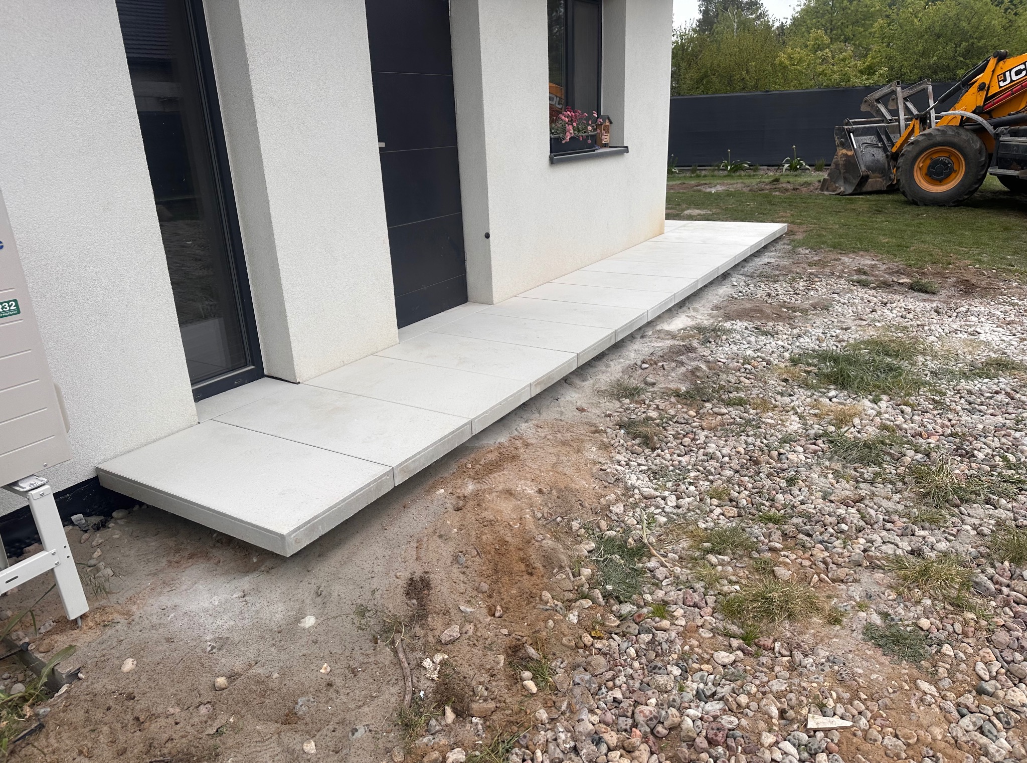 Ułożony chodnik z jasnych płyt przy nowoczesnym budynku z czarnymi drzwiami i oknem. W tle koparka JCB i teren z kamieniami.