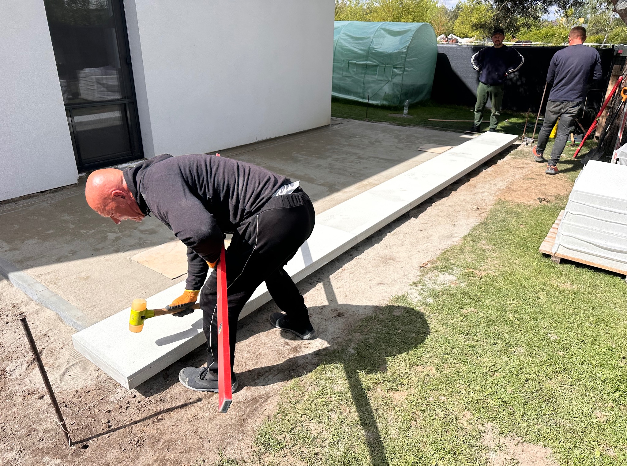 Pracownik z poziomnicą i młotkiem gumowym układa jasnoszare płyty na podwórku, w tle widoczni dwaj mężczyźni i szklarnia.