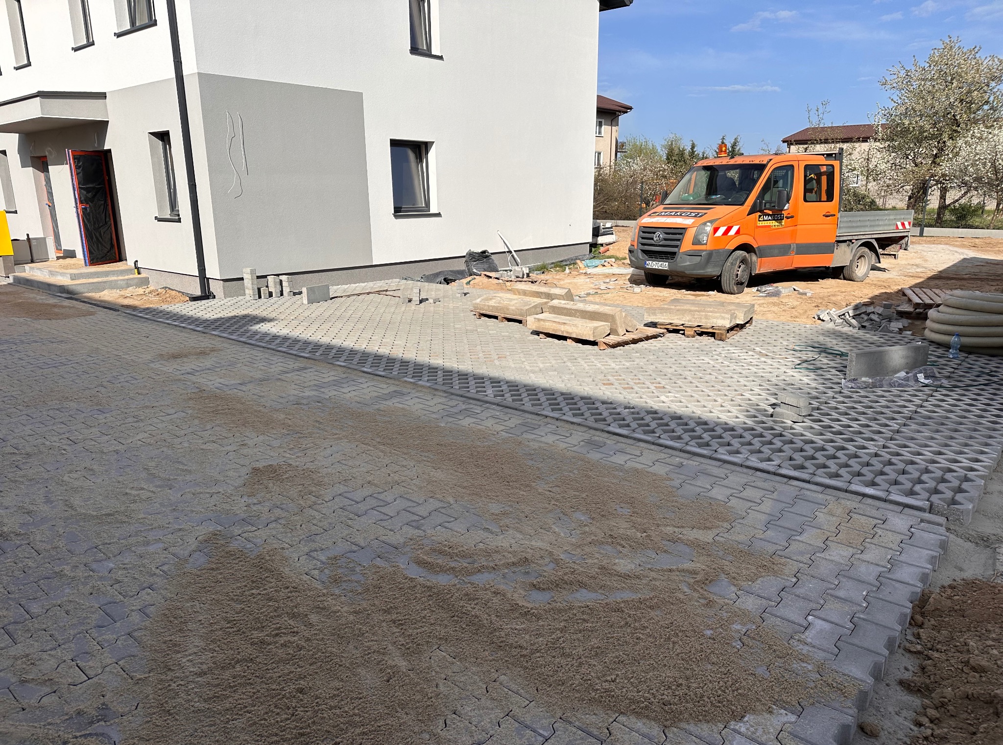 Przepuszczalna nawierzchnia z kostki ażurowej, częściowo zasypana piaskiem, przygotowana do dalszych prac. W tle budynek i pomarańczowy samochód dostawczy.
