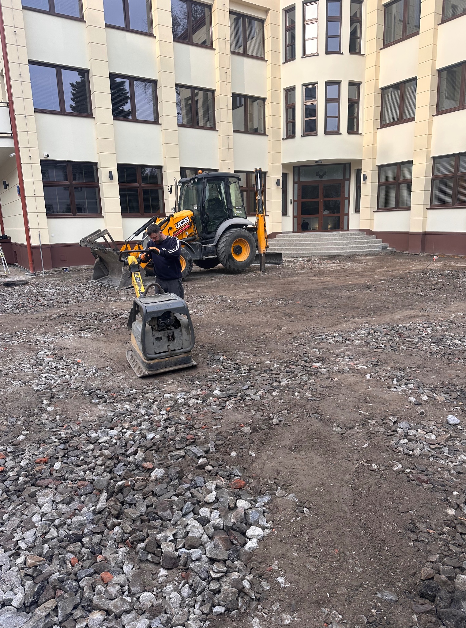 Pracownik ubija kamieniste podłoże zagęszczarką w pobliżu budynku, z koparką JCB w tle. Przygotowanie terenu pod budowę, widok z bliska.