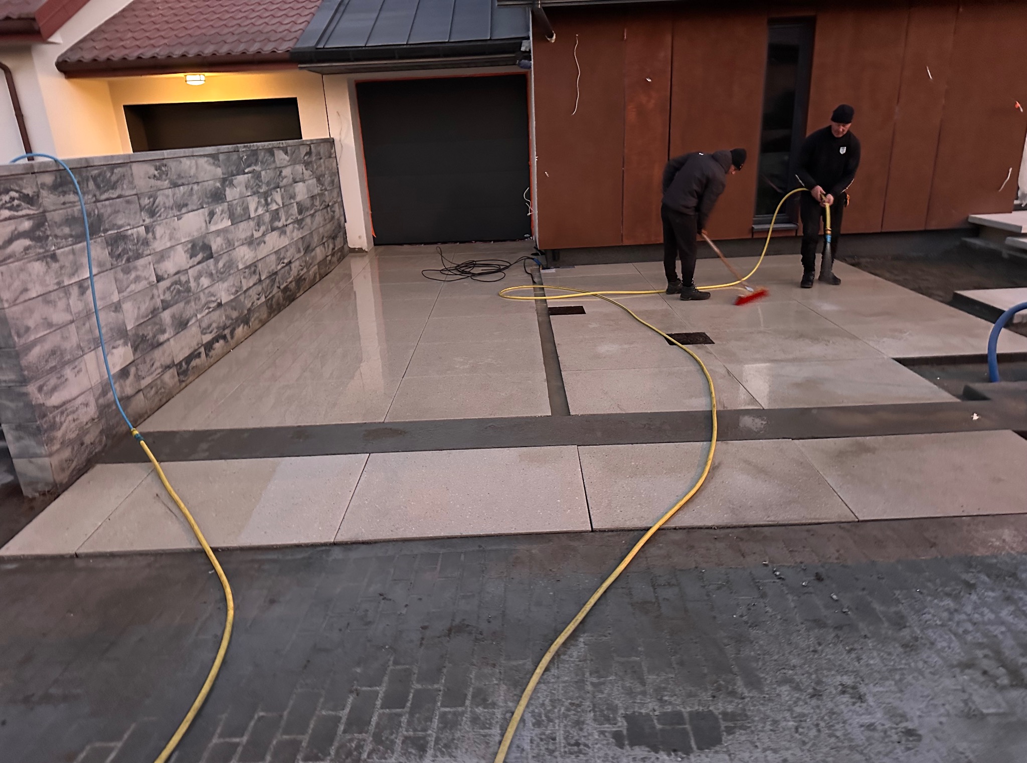 Układanie jasnej kostki brukowej z ciemnymi akcentami w Nowym Dworze Mazowieckim. Dwóch pracowników czyści powierzchnię po ułożeniu, używając wody i szczotki.