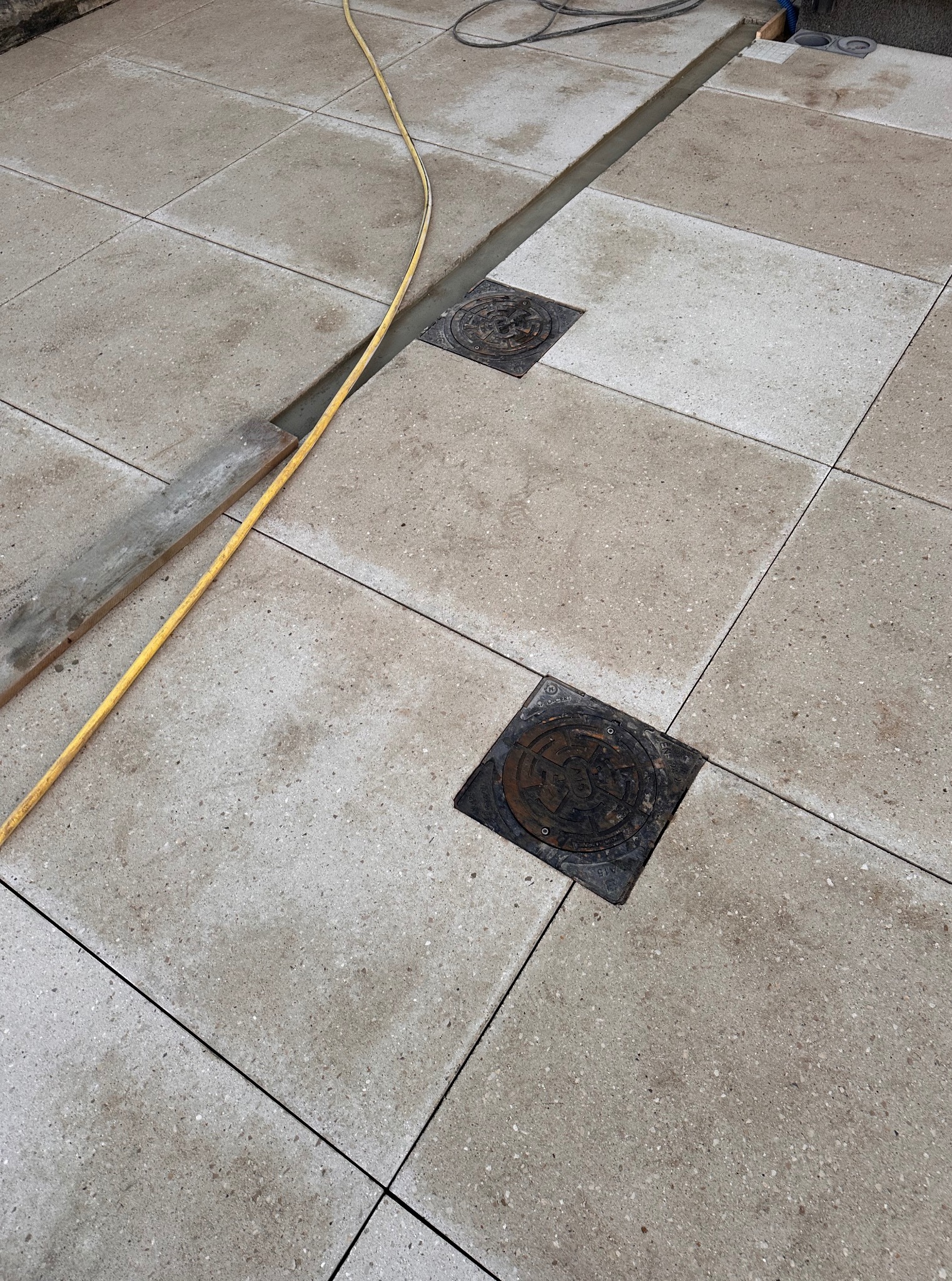 Ułożona, szara kostka brukowa z widocznymi studzienkami kanalizacyjnymi i żółtym wężem. Fragment deski widoczny w lewym górnym rogu kadru.