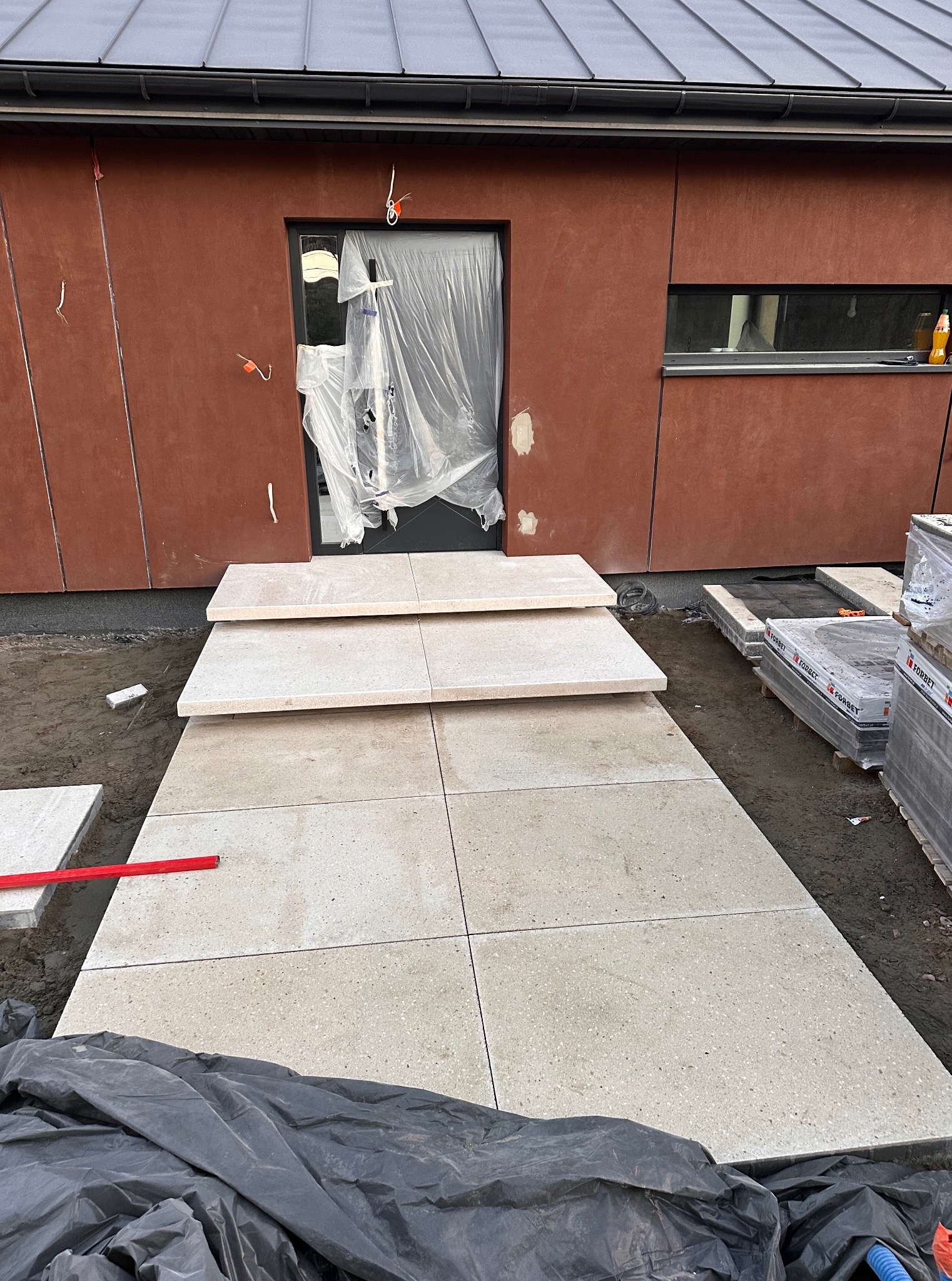 Jasne płyty chodnikowe prowadzą do drzwi wejściowych nowoczesnego domu. Widoczne palety z kostką i poziomica. Częściowo ukończona ścieżka z kamiennymi schodami.