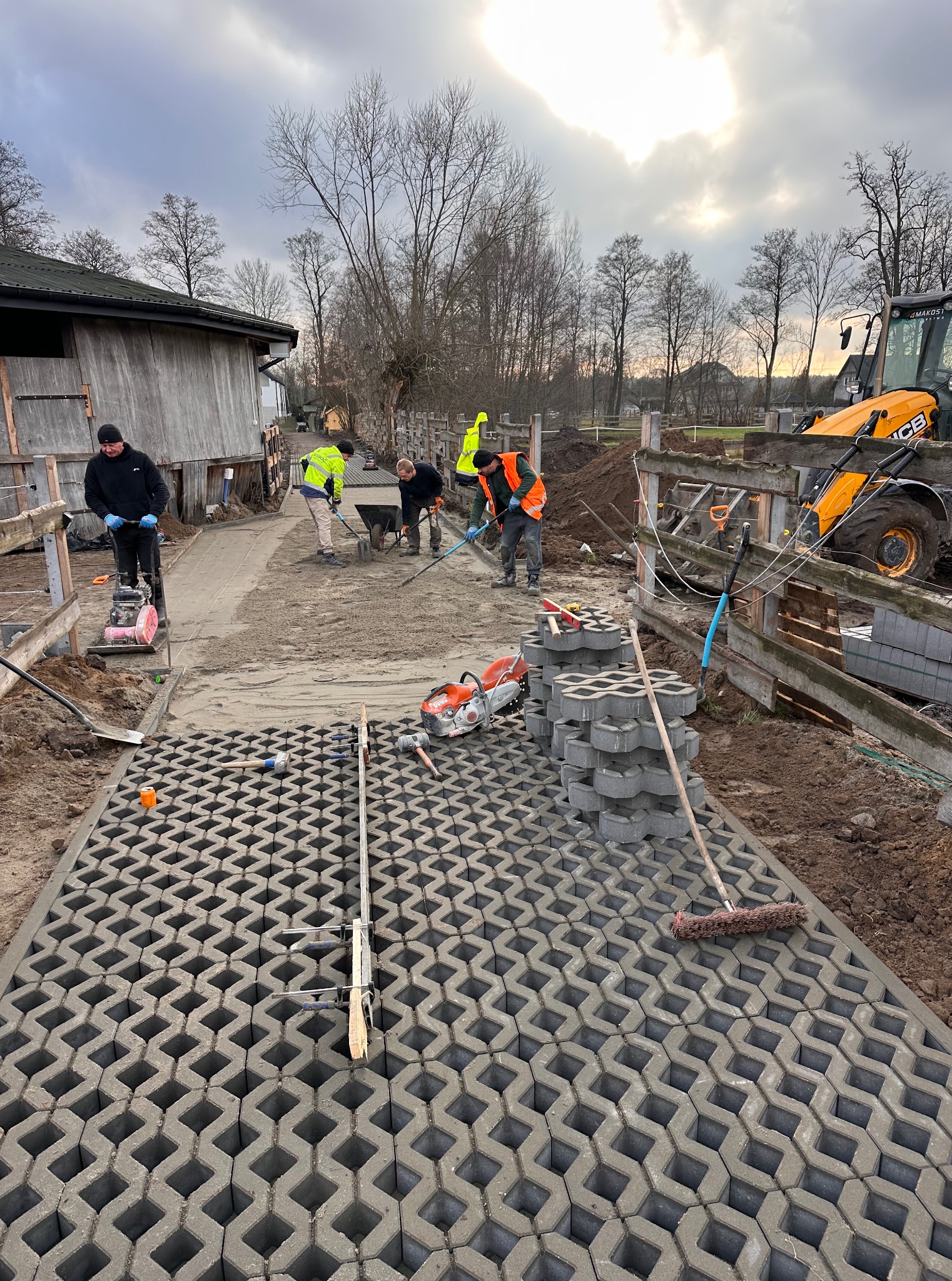 Ekipa budowlana w trakcie prac: część drogi wyłożona ażurową kostką, w tle maszyna budowlana i pracownicy wyrównujący teren. Pochmurne niebo.