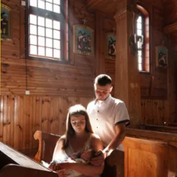 Rodzice z noworodkiem w drewnianym kościele. Matka trzyma dziecko na rękach, ojciec stoi za nią. Promienie słońca wpadają przez okno, oświetlając scenę.