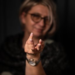 Beata Kołodziej - Kobieta z blond włosami i okularami, wyciąga dłoń z zegarkiem w kierunku widza, na ciemnym tle. Ujęcie z bliska, skupione na dłoni i zegarku.