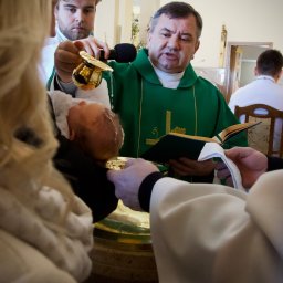 Kadr z ceremonii chrztu: ksiądz w zielonej szacie polewa wodą głowę dziecka, asystują rodzice. W tle wnętrze kościoła z delikatnym oświetleniem.