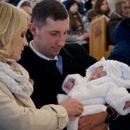 Rodzice z śpiącym niemowlęciem w białym ubranku podczas uroczystości w kościele. Mama delikatnie poprawia ubranko dziecka, w tle widoczni goście.