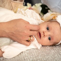 Urocze niemowlę w białej sukience i opasce z kokardą, leżące na miękkim dywanie, delikatnie dotykane przez dłoń z pierścionkiem. Kwiaty w tle.