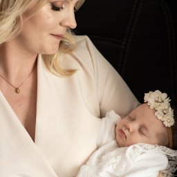 Portret matki z śpiącym noworodkiem w białej sukience i opasce z kwiatami. Delikatne oświetlenie, profil matki, ciemne tło. Ujęcie z bliska, skupienie na twarzach.