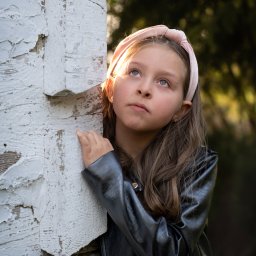 Beata Kołodziej - Dziewczynka w czarnej kurtce i różowej opasce, oparta o białą, chropowatą ścianę, spogląda w górę w promieniach słońca. Ujęcie z dołu, zieleń w tle.