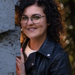 Beata Kołodziej - Portret uśmiechniętej kobiety w okularach i czarnej skórzanej kurtce, opierającej się o szarą, teksturowaną ścianę z widocznym złotym pierścionkiem i różowym lakierem do paznokci.