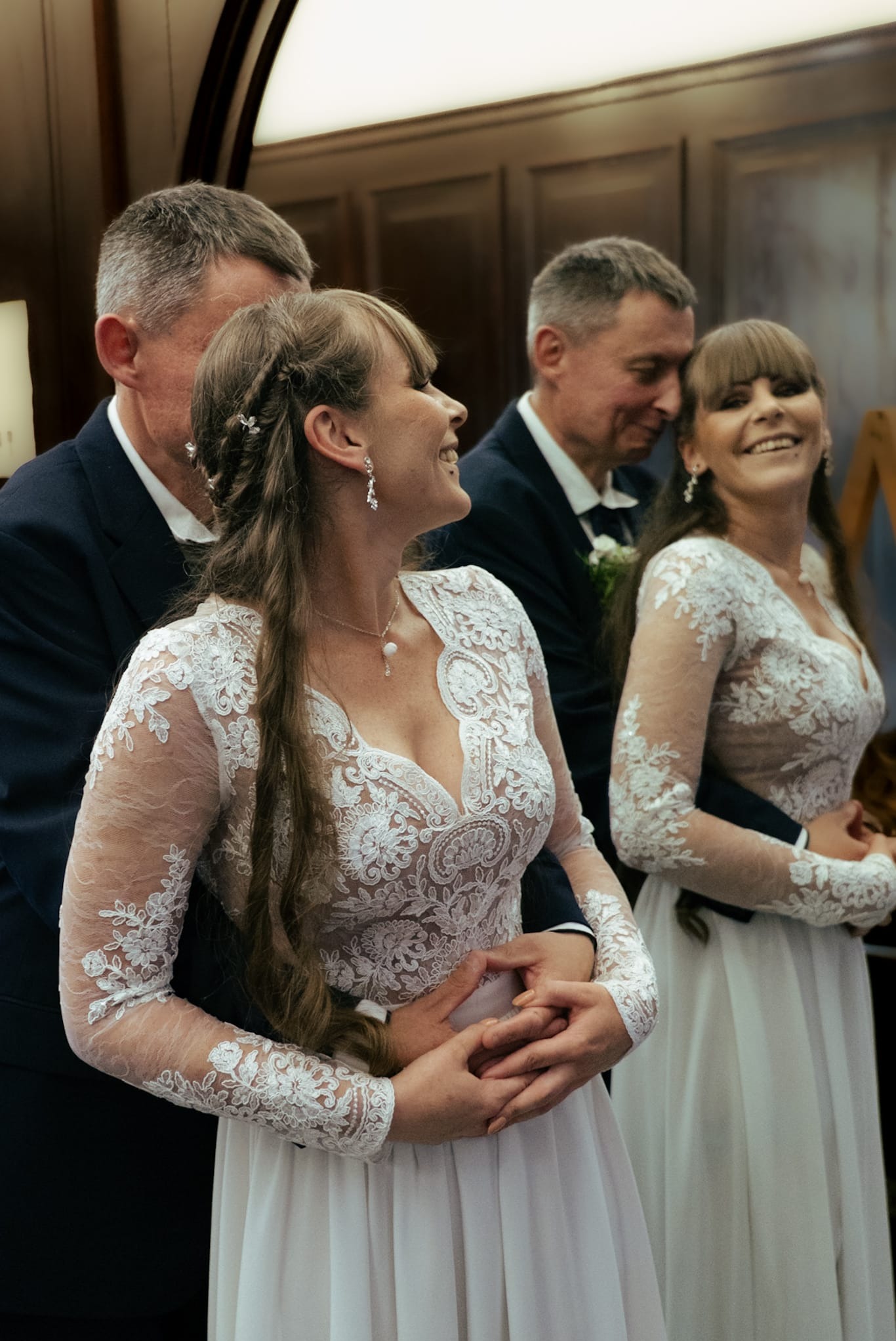 Uśmiechnięte panny młode w koronkowych sukniach, obejmowane przez mężczyzn w garniturach. Jasne wnętrze, subtelne oświetlenie, elegancka uroczystość.