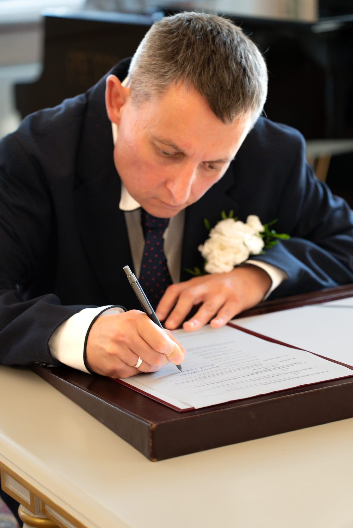 Elegancki mężczyzna w garniturze, z butonierką, podpisuje dokument na stole. Ujęcie z góry, skupione na dłoniach i dokumencie. W tle widoczny fortepian.