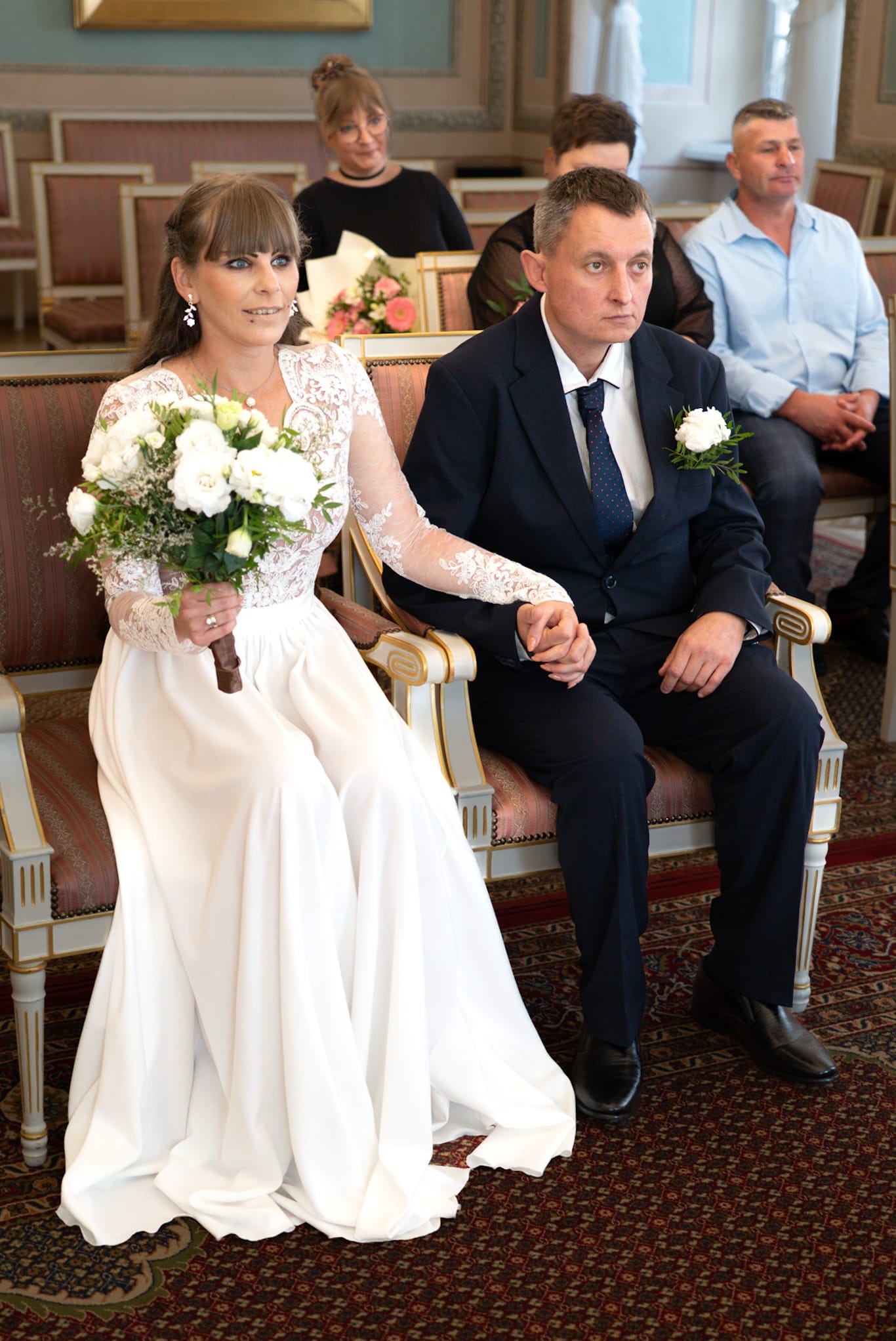 Para młoda siedzi obok siebie podczas ceremonii ślubnej, trzymając się za ręce. Panna młoda w białej sukni z bukietem, pan młody w granatowym garniturze z butonierką.