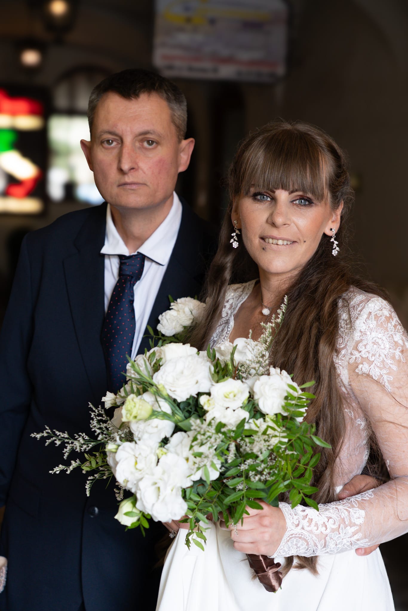 Para młoda: kobieta w koronkowej sukni ślubnej trzyma bukiet białych kwiatów, obok mężczyzna w granatowym garniturze. Ujęcie portretowe w ciemnym wnętrzu.