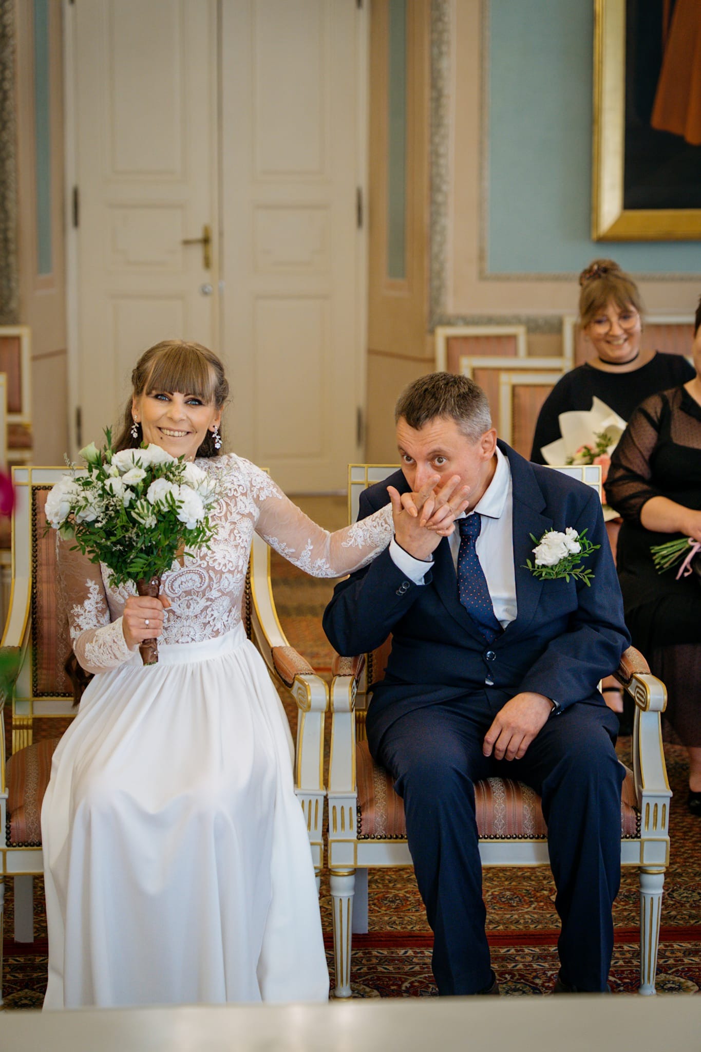Uśmiechnięta panna młoda z bukietem i pan młody całujący jej dłoń, siedzący na krzesłach podczas ceremonii ślubnej w jasnym wnętrzu.