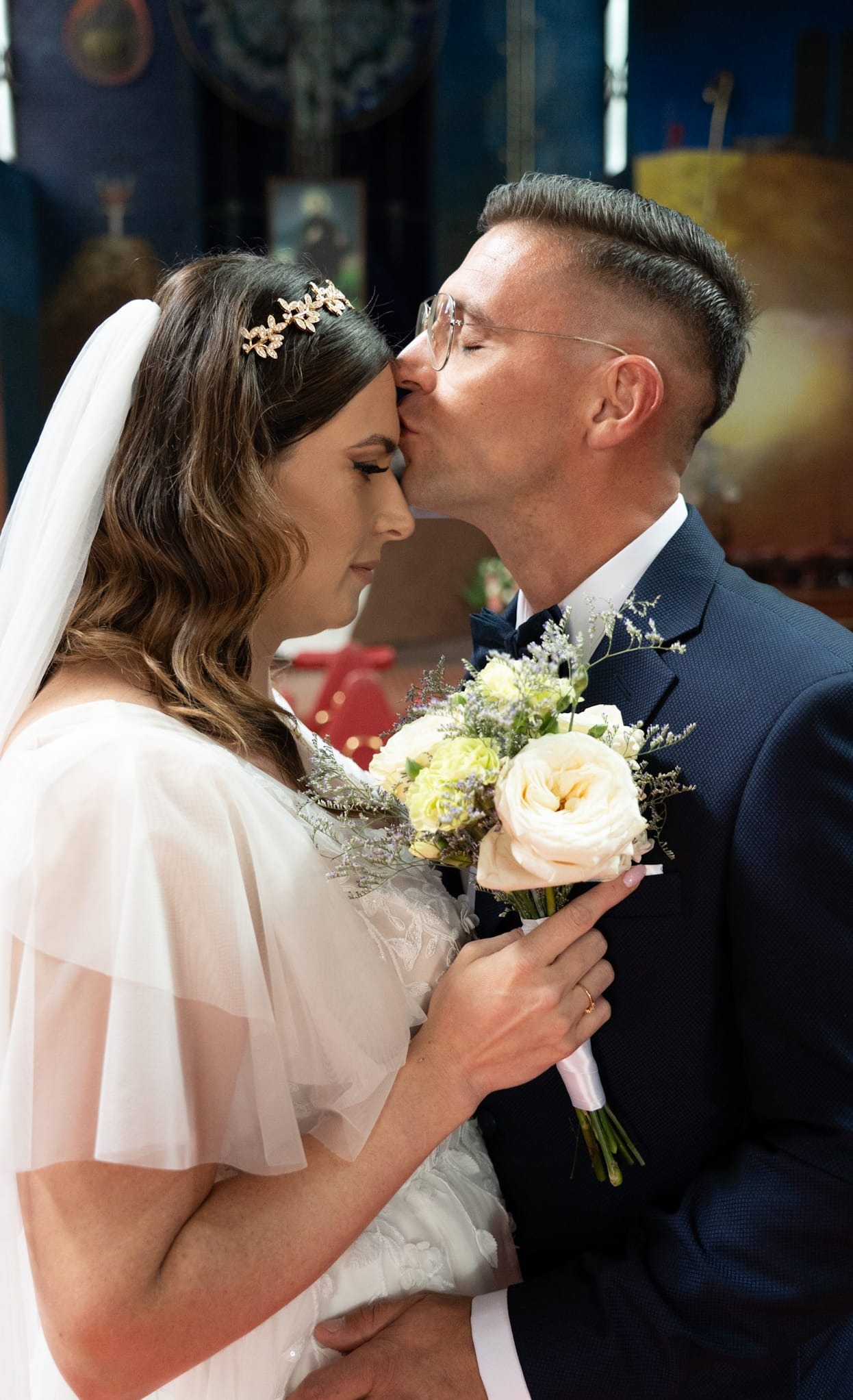 Panna młoda w sukni ślubnej z bukietem, oblubieniec całuje ją w czoło. Ujęcie w kościele, z widocznym fragmentem dekoracji ołtarza w tle.