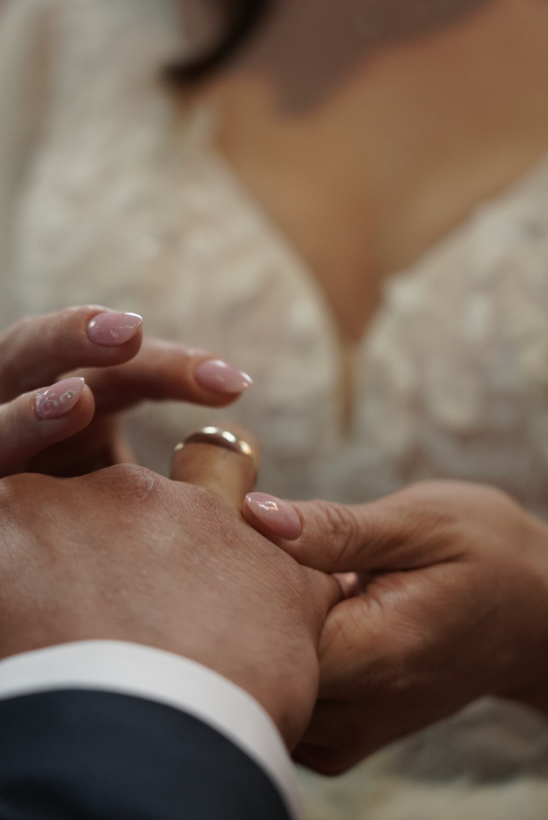 Moment zakładania obrączki podczas ceremonii ślubnej, zbliżenie na dłonie i złoty pierścionek, w tle suknia panny młodej z delikatnymi zdobieniami.